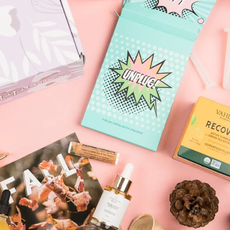 Cosmetic products and packaging on a pink surface with a textured blanket.