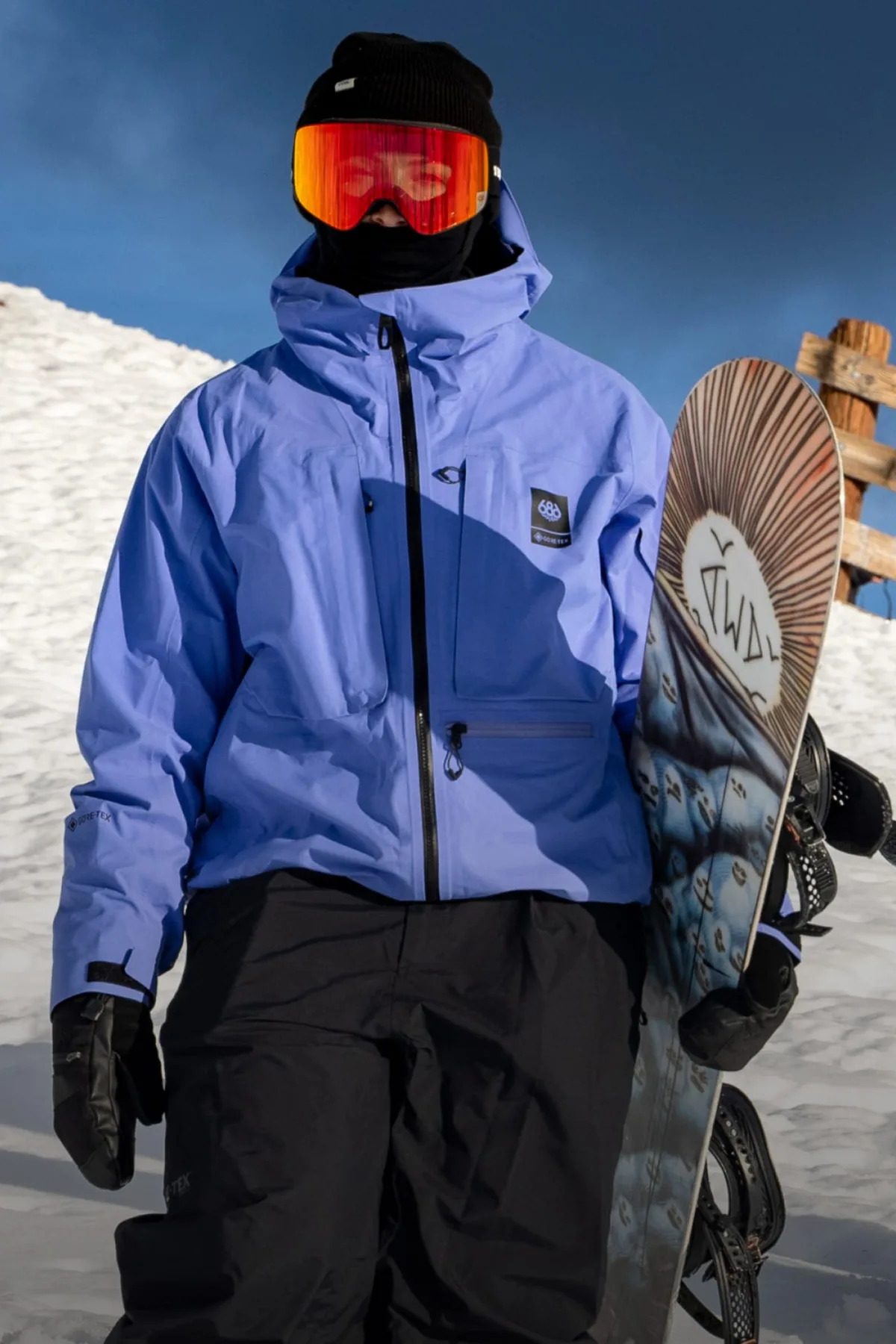 Snowboarder in a periwinkle 686 shell jacket and red goggles carrying a board on a snowy slope