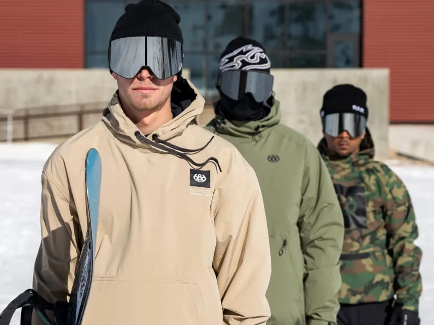 Three riders in 686 anoraks and goggles standing in the snow, one holding skis