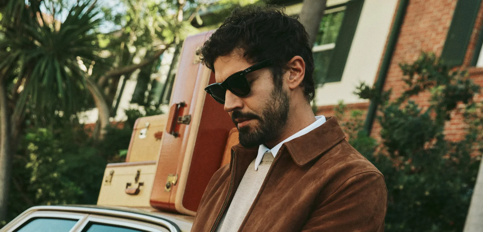 Man in sunglasses and a suede 7Diamonds jacket beside vintage suitcases on a car