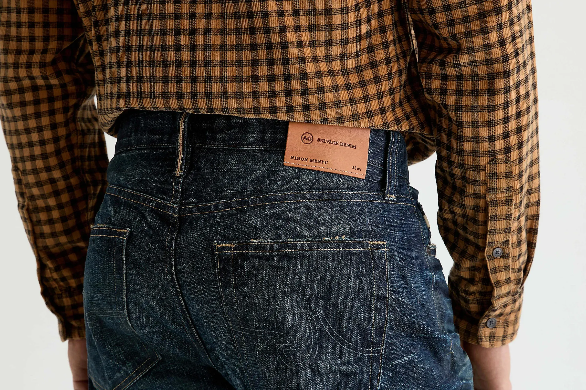 Back view of AG Jeans dark selvedge denim with a leather patch, worn with a gingham shirt