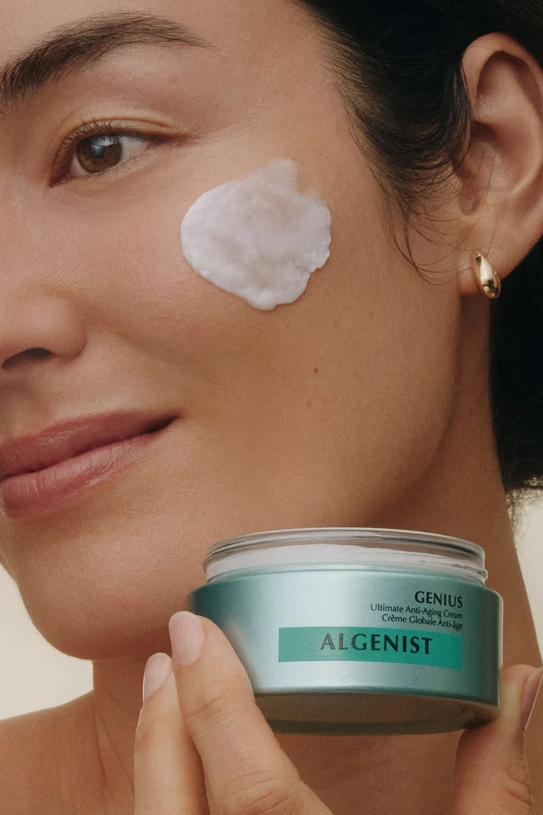 Woman holding a jar of Algenist GENIUS Ultimate Anti-Aging Cream, with cream on her cheek