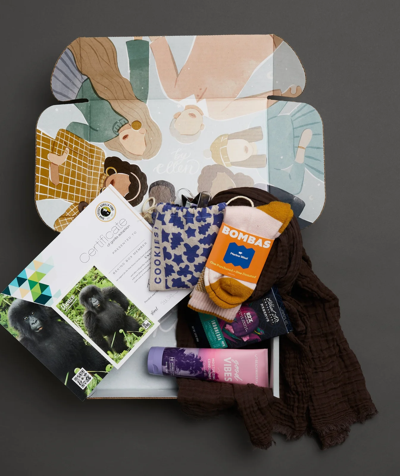 An open BE KIND. by ellen winter box with a gorilla adoption certificate, Bombas socks, cookie cutters, chocolate, conditioner and a scarf