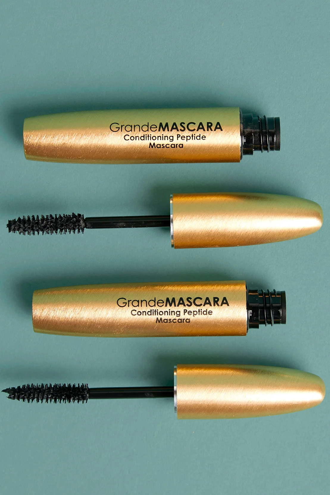 Two gold GrandeMASCARA conditioning mascaras laid on a teal backdrop