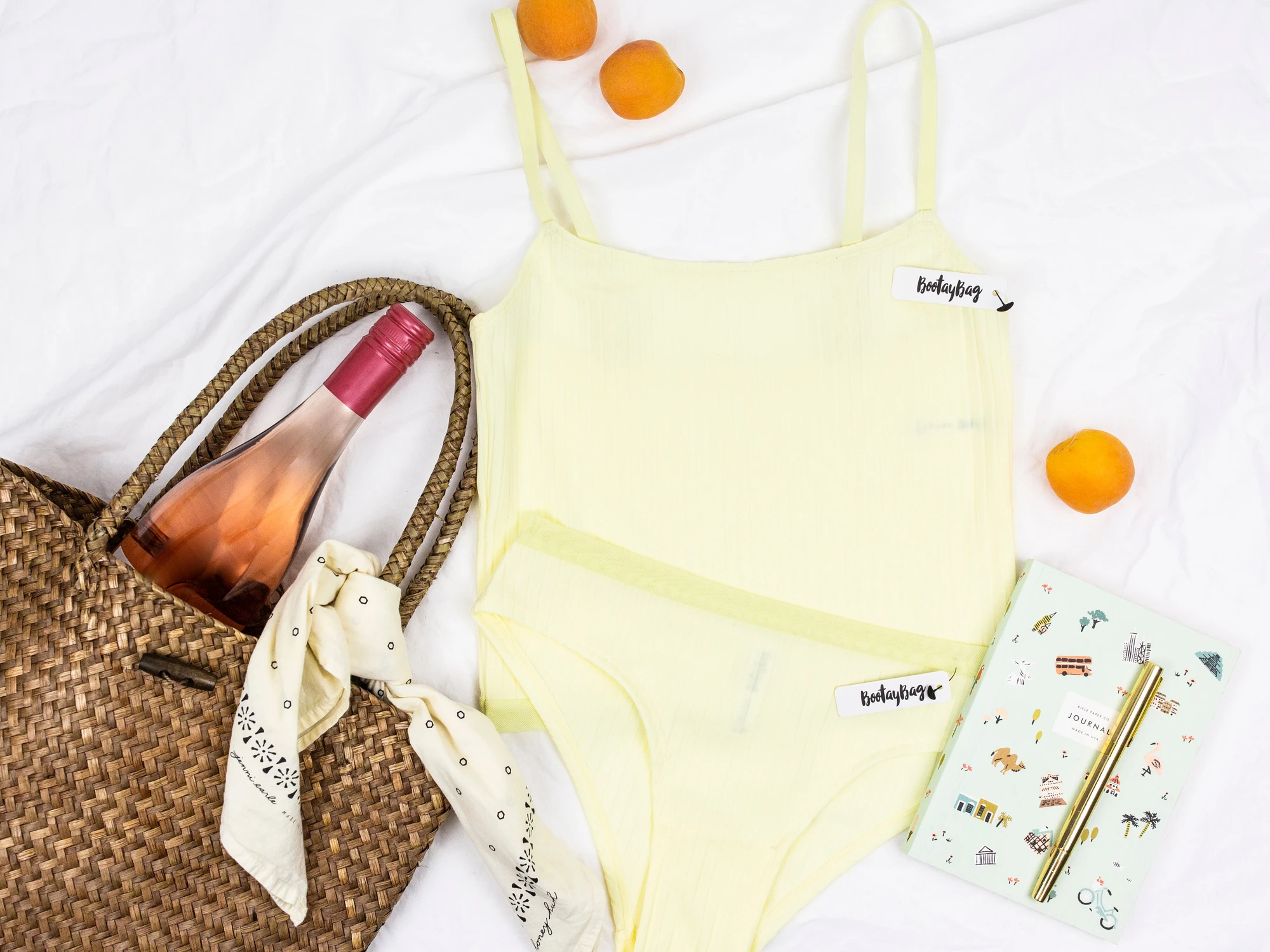 BootayBag pale yellow bodysuit and brief laid flat beside a straw bag, a bottle of rosé and a journal