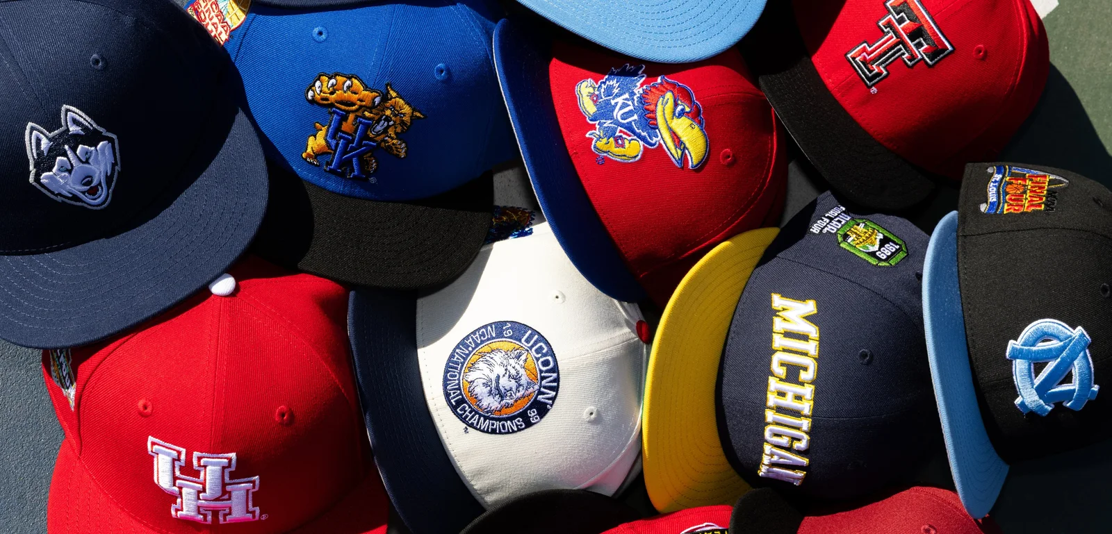 Pile of embroidered college snapback caps including UConn, Michigan, Kansas and North Carolina logos