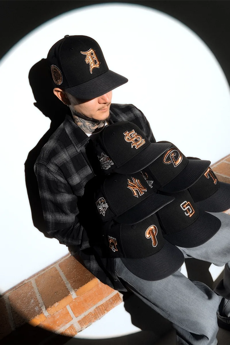 Man in a flannel shirt sitting on a brick ledge holding a stack of black Hat Club fitted caps