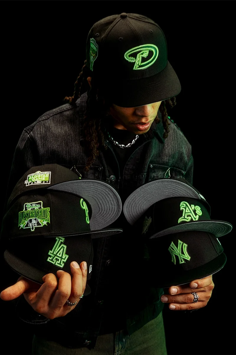Man holding black fitted caps with neon green team logos against a dark backdrop