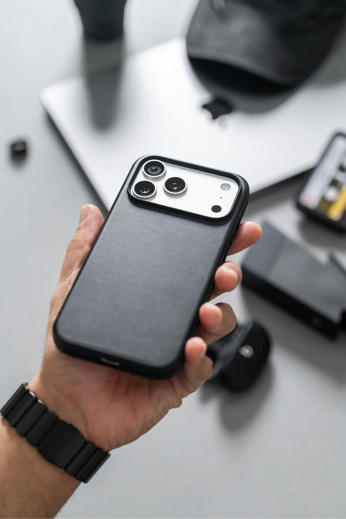 Hand holding an iPhone in a black Nomad case, with a laptop and accessories behind it