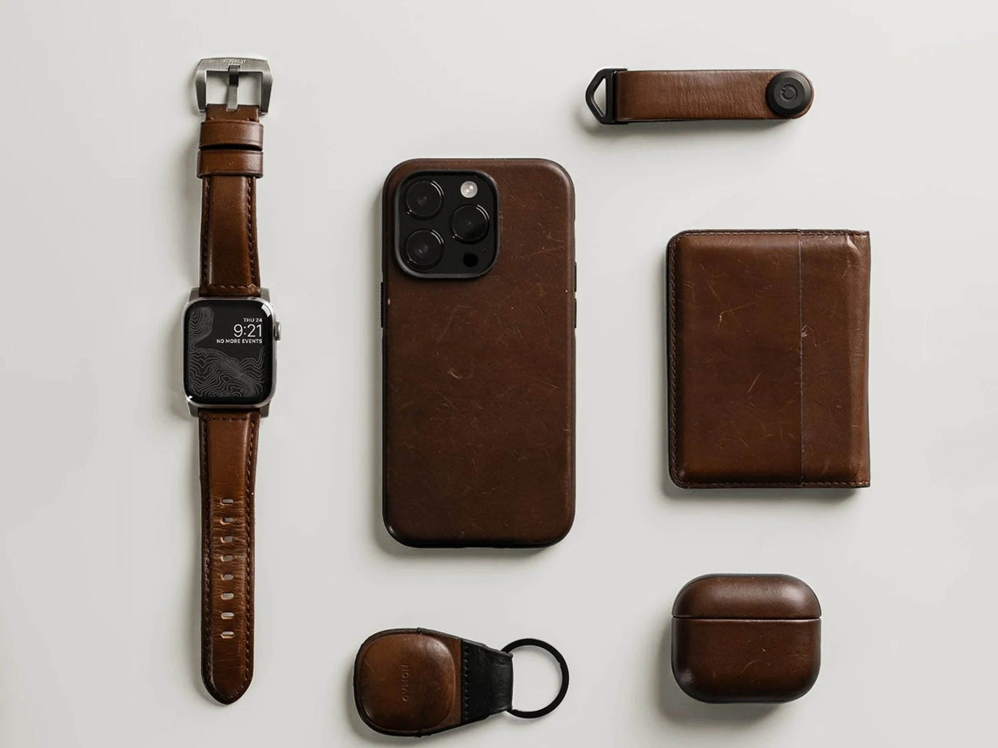 Flat lay of Nomad brown leather goods: watch band, iPhone case, wallet, key fob, keychain and AirPods case