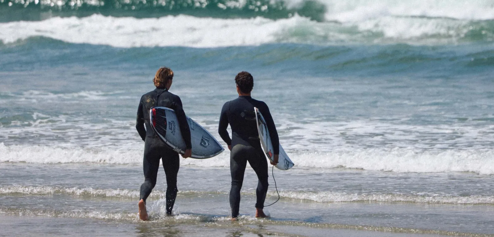 Two surfers in O’Neill wetsuits carrying boards into the waves