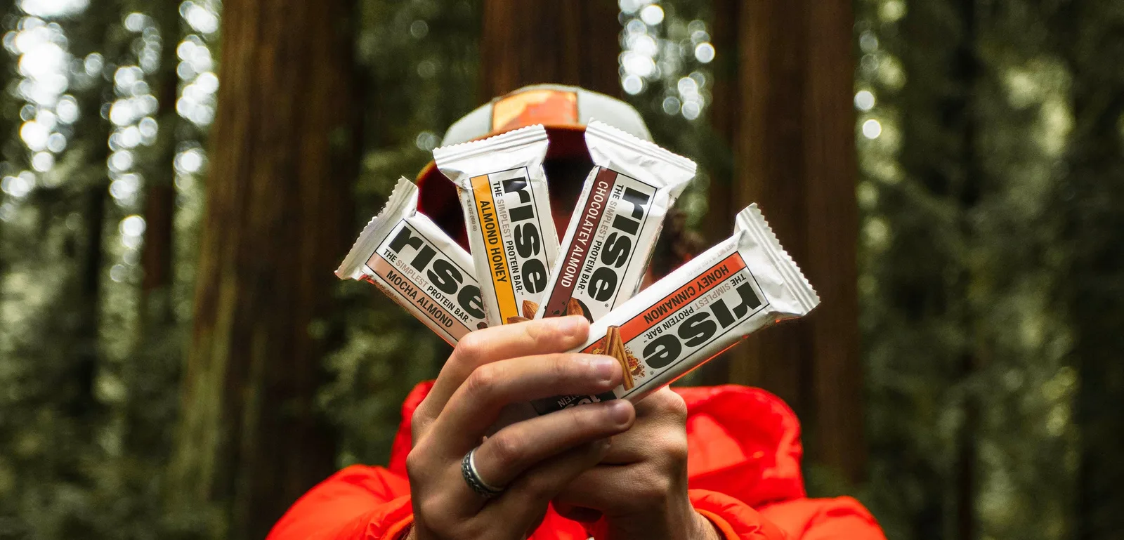 Hand holding four Rise protein bars in a redwood forest