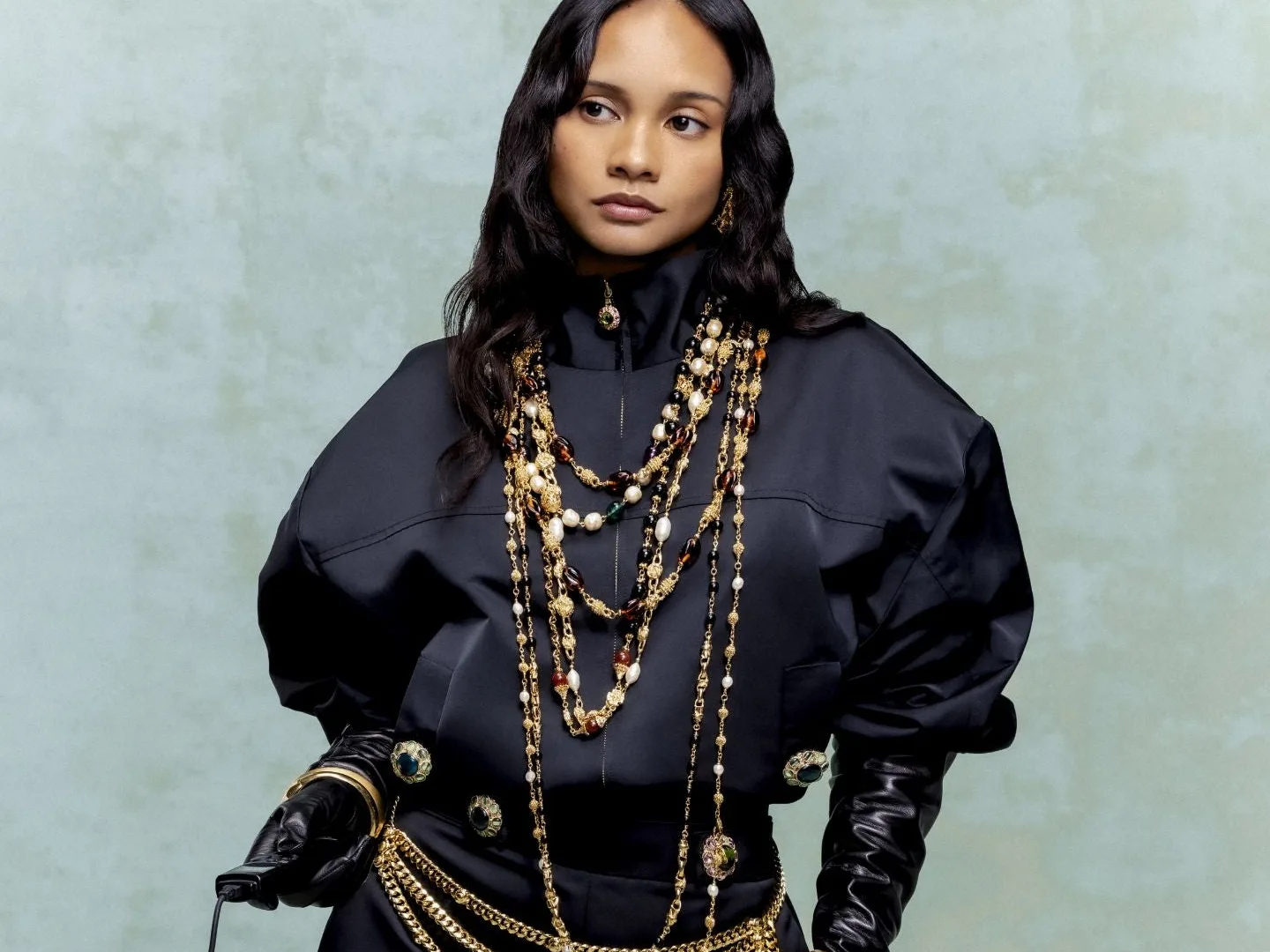 Model in a navy St. John jacket layered with gold and pearl chain necklaces on a sage backdrop