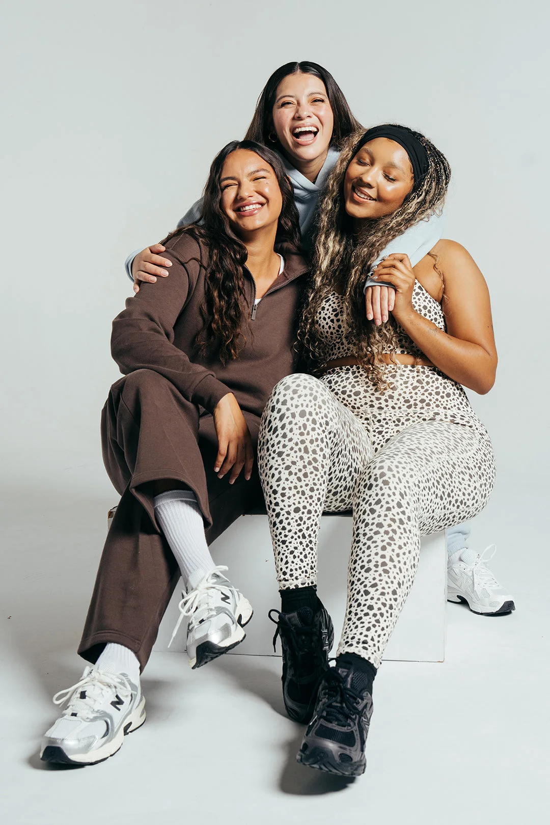 Three friends laughing on a white bench in sneakers and loungewear
